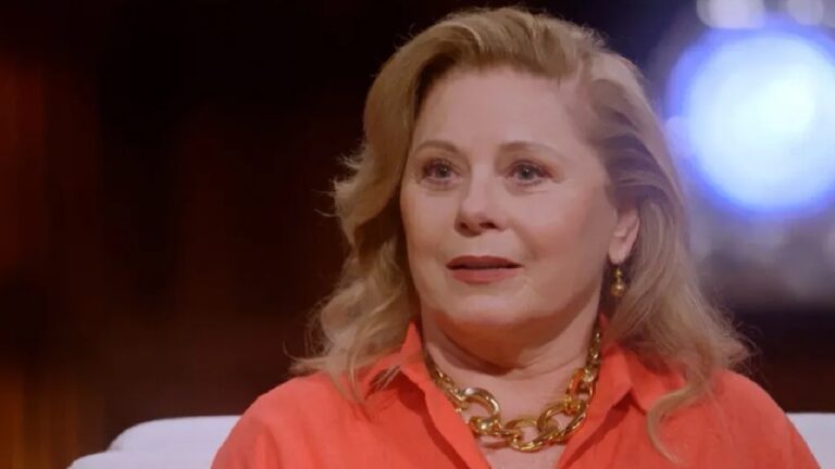 Mulher de blusa laranja e colar dourado sentada em estúdio iluminado com expressão atenta