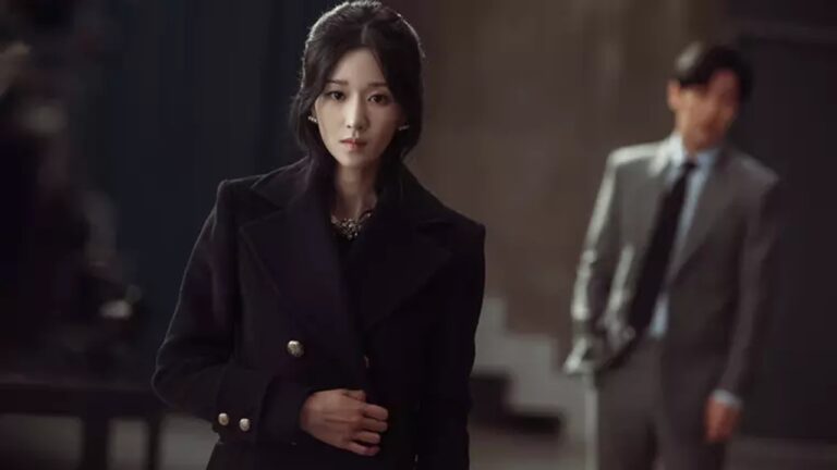 Mulher de casaco preto encara à frente enquanto homem de terno desfocado aparece ao fundo em ambiente interno elegante