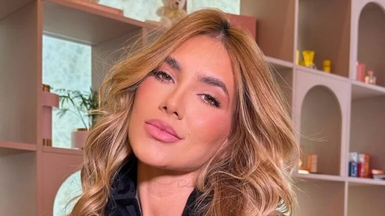 Mulher de cabelos loiros ondulados e maquiagem suave posa em ambiente interno bem iluminado, com prateleiras decoradas ao fundo