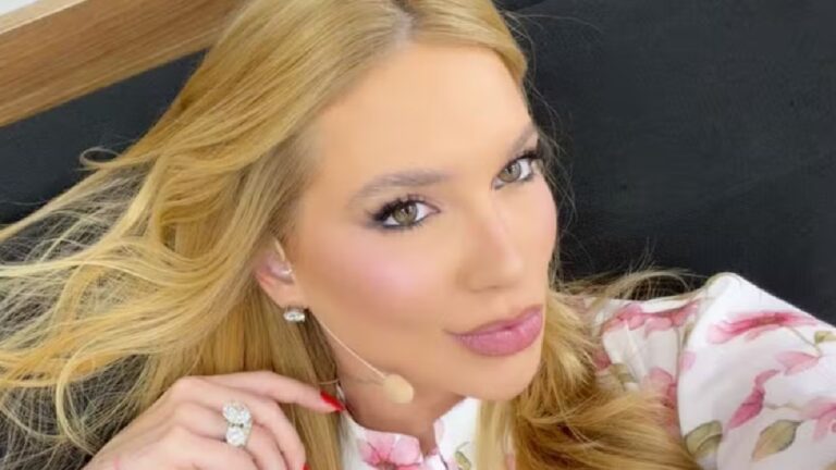 A imagem mostra uma mulher loira com cabelos soltos, maquiagem bem feita e batom rosado. Ela usa um microfone de orelha, indicando que provavelmente está participando de uma gravação ou programa. Está vestida com uma blusa branca estampada com flores cor-de-rosa e usa brincos e anel brilhante