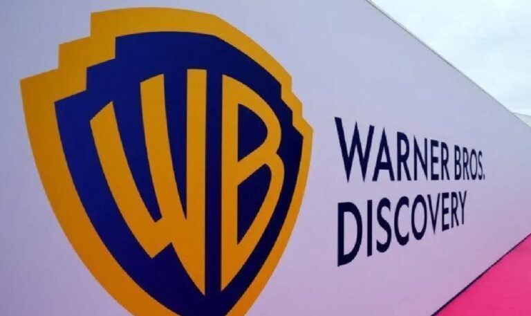 A imagem mostra o logotipo da Warner Bros. Discovery em um painel branco. À esquerda, está o icônico escudo dourado e azul com as iniciais “WB”, e à direita, o nome “Warner Bros. Discovery” aparece em letras maiúsculas pretas