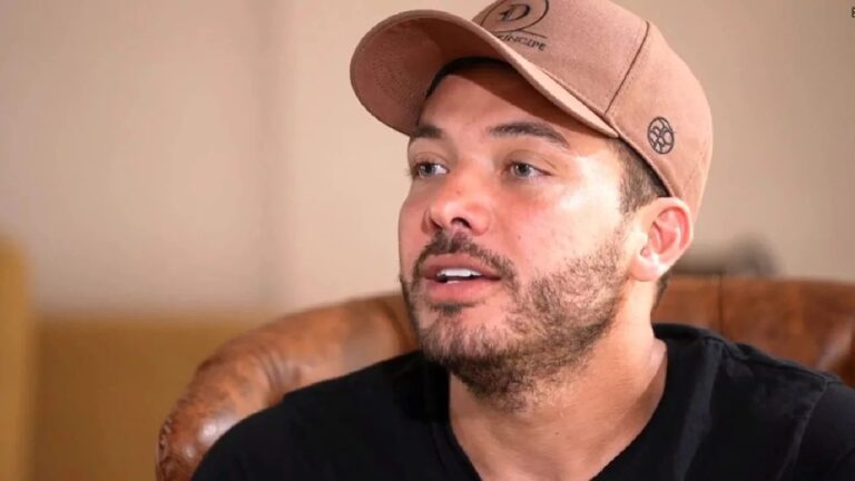 A imagem mostra um homem de barba curta e rala, usando boné marrom e camiseta preta, sentado em uma poltrona de couro. Ele olha ligeiramente para cima com expressão atenta, em um ambiente interno de tons neutros e iluminação suave