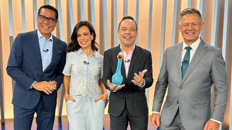 A imagem mostra Willian Leite, Fabiana Oliveira, Thiago Gardinali e Diógenes Lucca sorrindo em um estúdio iluminado, do Balanço Geral, com um fundo formado por painéis verticais em tons claros. Um deles segura um boneco azul em formato de ganso, sugerindo um momento leve e descontraído durante o programa da Record