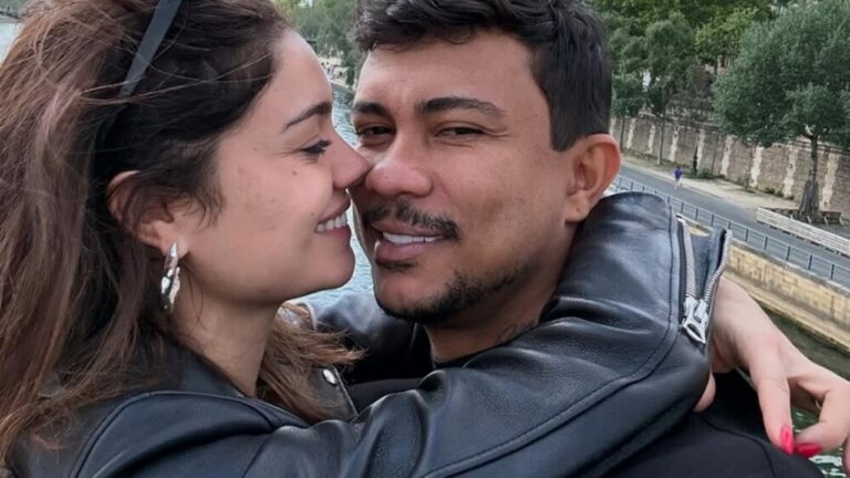 A imagem mostra um casal em um momento carinhoso, abraçados e sorrindo um para o outro. Ambos estão usando jaquetas de couro pretas. O cenário ao fundo parece ser uma cidade com um rio e árvores, sugerindo que a foto foi tirada ao ar livre, em um ambiente urbano e romântico