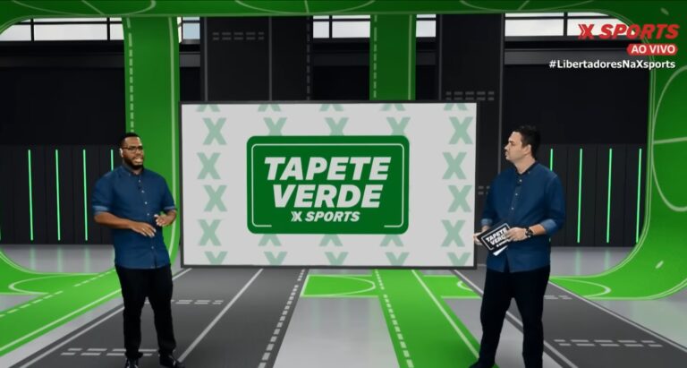 A imagem mostra dois apresentadores em um estúdio esportivo com o logotipo “Tapete Verde X Sports” ao fundo. O cenário é moderno, com tons de verde e cinza remetendo a um campo de futebol. Ambos vestem camisas azul-escuras e conversam durante a transmissão ao vivo, conforme indicado pelo selo “AO VIVO” no canto superior direito