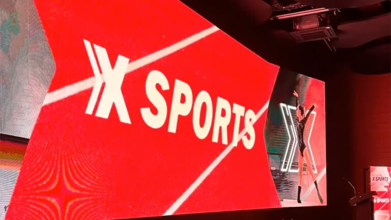 Telão vermelho exibindo o logo “X Sports” ocupa a maior parte da cena, com imagem lateral de atleta posando em traje esportivo; ambiente interno de evento com iluminação de palco e púlpito ao lado