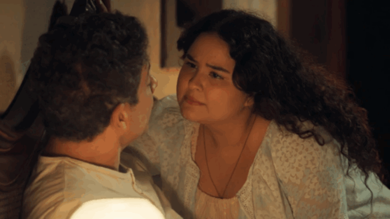 Cena mostra duas pessoas em um momento de proximidade e emoção. A mulher, com expressão séria e olhar intenso, está inclinada em direção ao homem que está sentado, sugerindo uma conversa importante ou tensa. O ambiente tem iluminação quente e acolhedora, típica de um cenário doméstico