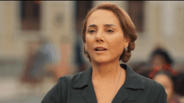 Mulher de expressão séria aparece em ambiente externo com construções históricas ao fundo, vestindo roupa escura e colar discreto, em cena de tom dramático