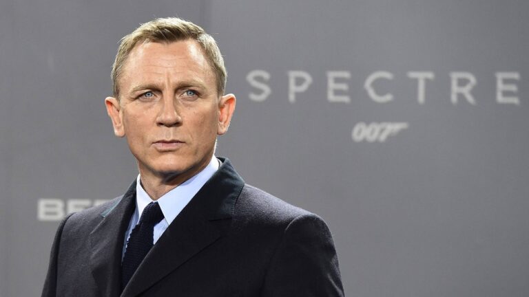 Homem de terno escuro e expressão séria posa diante de painel cinza com o logotipo do filme Spectre 007 ao fundo
