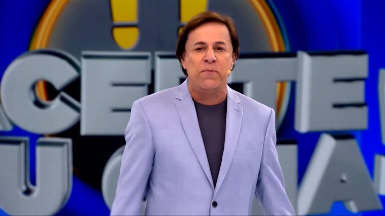 A imagem mostra Tom Cavalcante em um estúdio colorido, vestindo um paletó azul-claro e camiseta cinza. Ele usa microfone de lapela e está diante de um painel com letras grandes que fazem parte do logotipo do programa Acerte ou Caia!. O cenário tem tons de azul e iluminação intensa, típicos de programas de auditório