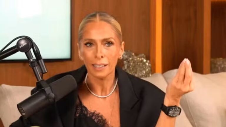 Mulher de cabelos presos para trás, vestindo blazer escuro e colar, fala em um estúdio de podcast enquanto gesticula com a mão, com um microfone profissional ao lado e cenário interno moderno ao fundo.