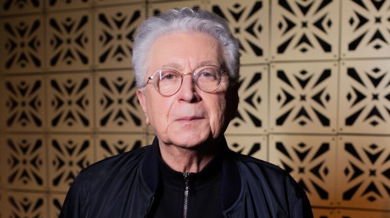 Homem idoso olha diretamente para a câmera em um retrato feito em ambiente interno. Ele tem cabelos curtos e brancos, usa óculos de armação redonda e veste jaqueta escura sobre roupa preta. Ao fundo, uma parede com padrão geométrico vazado cria textura visual, com iluminação suave que destaca o rosto e transmite um tom sóbrio e formal.