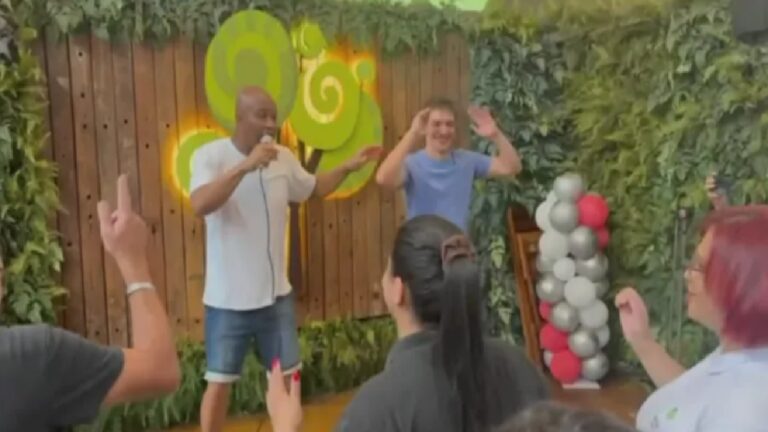 Dois homens se apresentam em um pequeno palco montado ao ar livre. Um deles segura um microfone e fala ao público, enquanto o outro, ao lado, levanta as mãos sorrindo. À frente do palco, pessoas participam com os braços erguidos. O cenário tem parede de madeira, plantas decorativas, um letreiro verde iluminado e uma coluna de balões em tons de branco, cinza e vermelho, sugerindo um evento descontraído ou comemorativo.