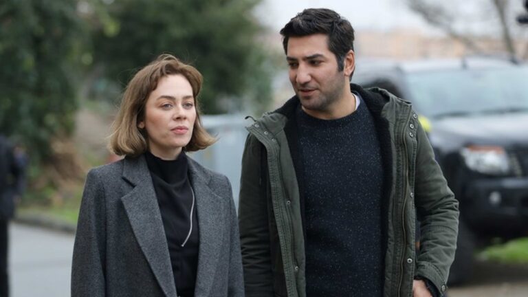 Duas pessoas caminhando lado a lado em uma rua ao ar livre em dia nublado. A mulher, à esquerda, usa casaco cinza e blusa preta, com expressão neutra. O homem, à direita, veste jaqueta verde e suéter escuro, olhando para ela enquanto conversam. Ao fundo, árvores desfocadas e carros estacionados completam a cena urbana.