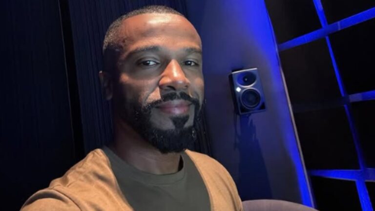 Homem com barba e camiseta marrom sorri levemente em estúdio de gravação iluminado por luz azul