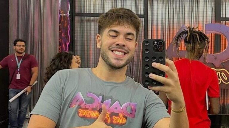 Homem sorridente tira selfie com celular em frente a espelho enquanto faz sinal de positivo usando camiseta cinza com estampa colorida, em ambiente interno com outras pessoas ao fundo