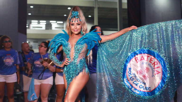 Ana Beatriz Godoi, Rainha de Bateria, usando fantasia azul com penas e lantejoulas, segurando a bandeira da Rosas de Ouro