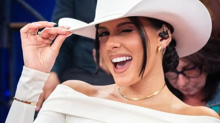 Mulher sorridente veste chapéu branco de estilo country e pisca um olho enquanto segura a aba; ela usa blusa clara de ombros à mostra, acessórios dourados e tem um ponto eletrônico na orelha, em um ambiente interno com pessoas desfocadas ao fundo.