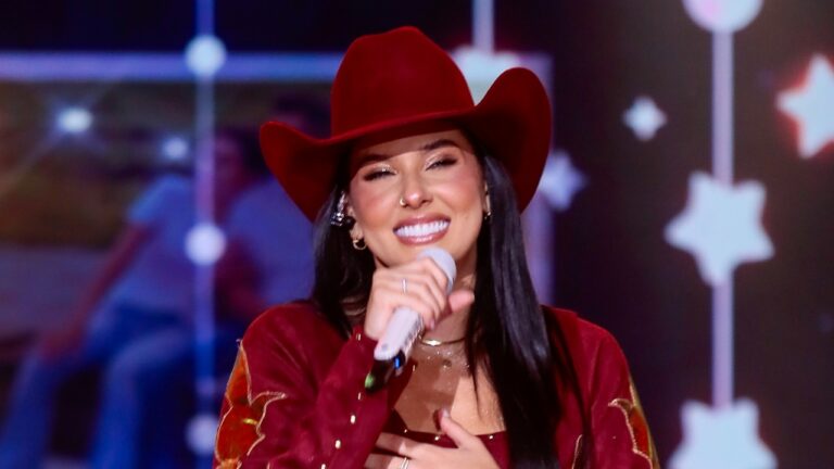 A imagem mostra Ana Castela, uma mulher sorridente, usando um chapéu de cowboy vermelho e segurando um microfone, enquanto se apresenta em um palco iluminado. O fundo tem um visual decorativo com estrelas desfocadas, criando um clima festivo e musical. Ela parece estar cantando, transmitindo alegria e carisma durante a performance