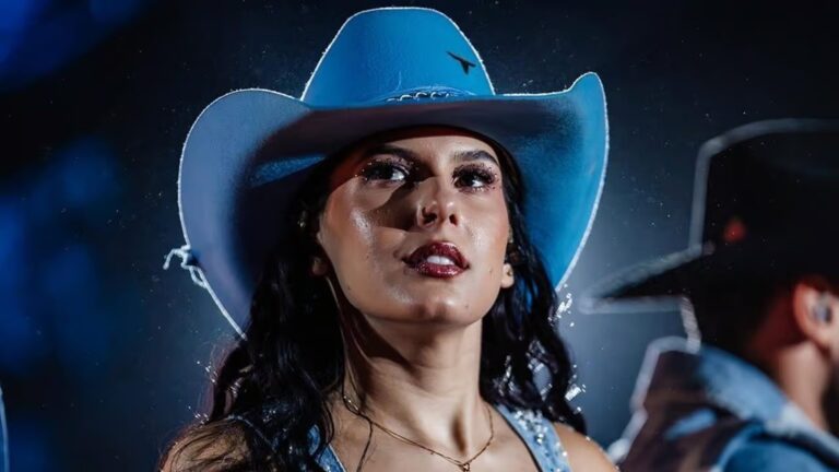 A imagem mostra a cantora Ana Castela usando chapéu de cowboy azul-claro, com maquiagem brilhante e expressão confiante, sob luzes de palco. Ela veste uma roupa no mesmo tom de azul e parece estar em uma apresentação musical noturna, com outro músico desfocado ao fundo. O ambiente tem atmosfera de show, com iluminação cênica e partículas visíveis no ar