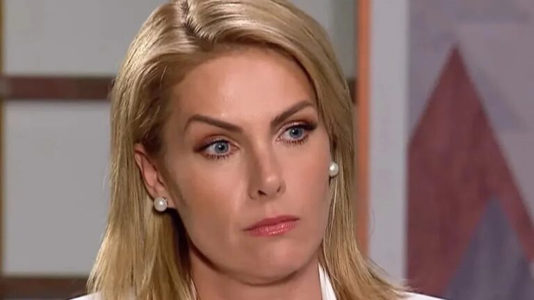 Close da face de uma mulher adulta de cabelos loiros lisos, usando brincos de pérola e roupa clara. Ela olha levemente para o lado, com expressão séria e concentrada. O fundo é desfocado, com tons neutros e um elemento geométrico, sugerindo um ambiente interno de estúdio ou entrevista.