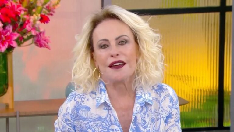 Apresentadora de cabelos loiros cacheados, usando camisa estampada em azul e branco, aparece sorrindo em um estúdio iluminado. Ao fundo, há flores vibrantes em tons de rosa e elementos decorativos com luz amarelada, criando um clima acolhedor de programa matinal.