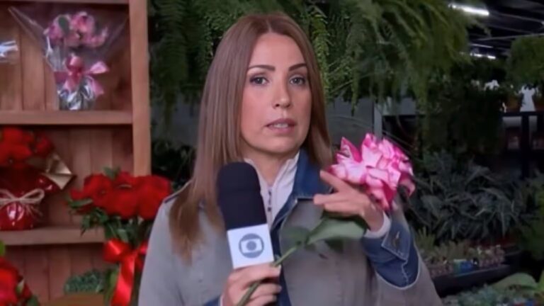 Repórter segura um microfone e uma flor rosa enquanto fala em frente a um estande repleto de arranjos florais; atrás dela há plantas verdes penduradas e prateleiras com buquês coloridos.
