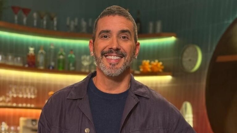 Homem sorridente, com barba curta grisalha e cabelo também grisalho, aparece em ambiente interno iluminado. Ele usa camiseta escura e camisa casual aberta. Ao fundo, há prateleiras com garrafas e copos, além de iluminação colorida que cria clima de estúdio ou bar moderno.