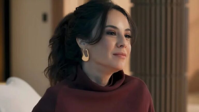 Mulher adulta com cabelo escuro preso em um rabo de cavalo volumoso, usando blusa de gola alta em tom vinho e brinco dourado geométrico. Ela aparece em ambiente interno elegante, com iluminação suave e elementos arquitetônicos desfocados ao fundo, mantendo uma expressão serena.