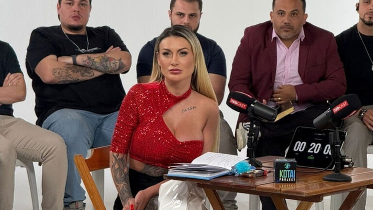 Andressa Urach veste top vermelho de paetê e olha para frente em entrevista de podcast com microfones e homens sentados atrás