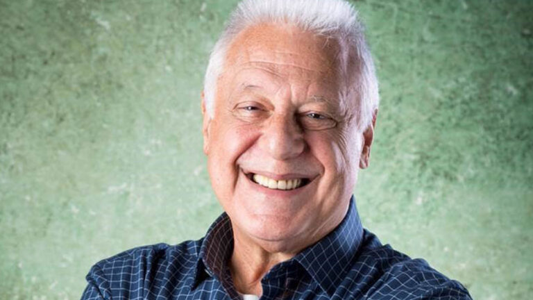 Antonio Fagundes sorrindo com cabelo branco, camisa xadrez azul e braços cruzados em foto com fundo verde texturizado