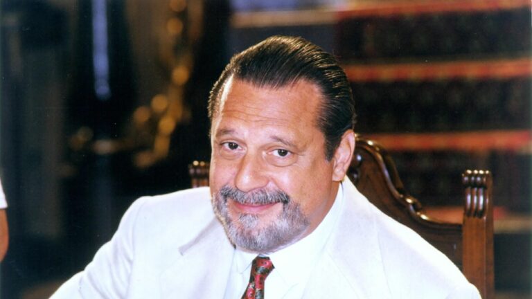 Homem de meia-idade, com barba e bigode grisalhos, cabelo penteado para trás e expressão sorridente, veste terno branco com gravata colorida; está sentado em uma cadeira de madeira trabalhada, com um ambiente interno desfocado ao fundo.