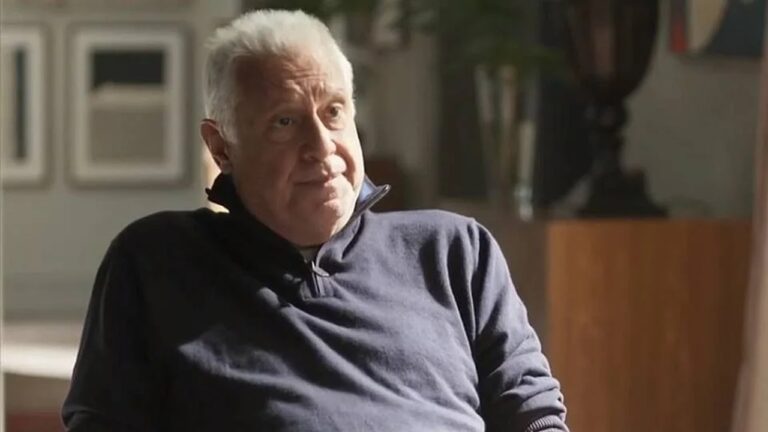 Homem idoso de cabelos brancos e expressão pensativa está sentado, vestindo um suéter azul escuro com zíper no colarinho, em um ambiente interno iluminado naturalmente, com móveis e quadros desfocados ao fundo.