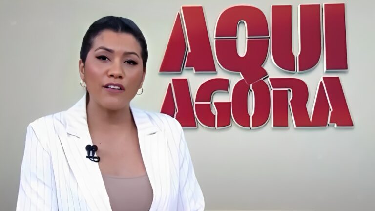 A imagem mostra Daniele Brandi, uma apresentadora de televisão vestida com um blazer branco e uma blusa bege, em frente a um fundo claro. Atrás dela, há um grande logotipo vermelho com as palavras Aqui Agora, sugerindo que ela está em um cenário de programa jornalístico. Ela usa um microfone de lapela e tem uma expressão neutra e profissional