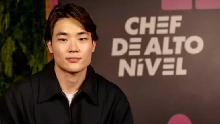 Homem jovem com aparência asiática posa em frente a um painel de evento com o texto “CHEF DE ALTO NÍVEL” ao fundo. Ele tem cabelo curto e escuro, penteado de lado, expressão neutra e veste uma jaqueta preta. O cenário é de estúdio ou coletiva de imprensa, com fundo escuro e iluminação profissional.