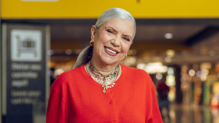 Astrid Fontenelle, com cabelos grisalhos e longos, em um suéter de tricô vermelho, sorrindo e usando colares de contas e pingentes, em um ambiente interno de aeroporto