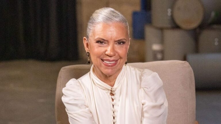 Mulher de cabelos grisalhos presos para trás, usando blusa clara de mangas bufantes, sorri enquanto está sentada em uma poltrona em ambiente interno com fundo desfocado.