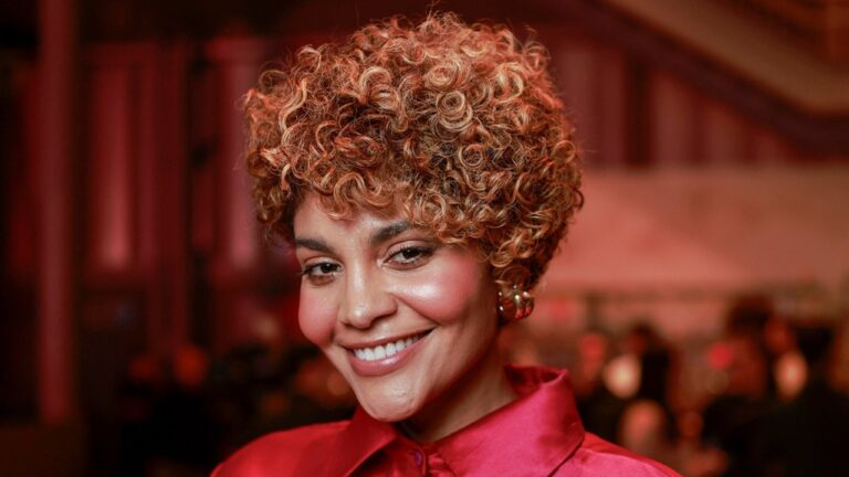 Mulher sorridente com cabelo curto e cacheado, em tons loiros, usa camisa de tecido brilhante em cor vermelha. Ela está em um ambiente interno com iluminação avermelhada e pessoas desfocadas ao fundo, sugerindo um evento social ou festa