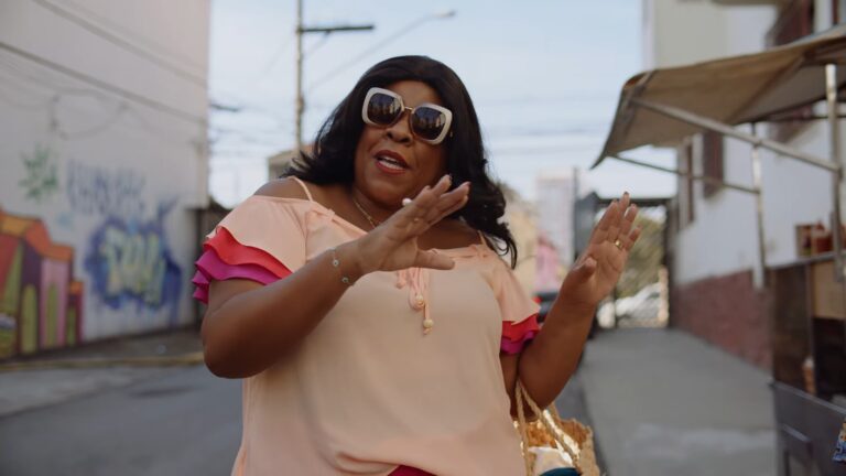 A imagem mostra uma mulher em uma rua urbana, usando óculos de sol grandes e blusa em tons de pêssego e rosa com babados nas mangas. Ela gesticula com as mãos, aparentando estar em uma conversa animada. Ao fundo, há muros grafitados e barracas de rua, compondo um cenário descontraído e cotidiano
