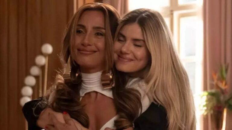 Duas mulheres adultas se abraçam em um ambiente interno bem iluminado. A mulher em primeiro plano tem cabelos longos e castanhos, usa blusa clara com decote alto e expressão emocionada, com os olhos marejados. A outra, de cabelos longos e loiros, abraça-a por trás com os olhos fechados e um leve sorriso, transmitindo carinho e apoio. Ao fundo, há uma janela com luz natural e elementos decorativos, sugerindo um ambiente doméstico ou de cena dramática.