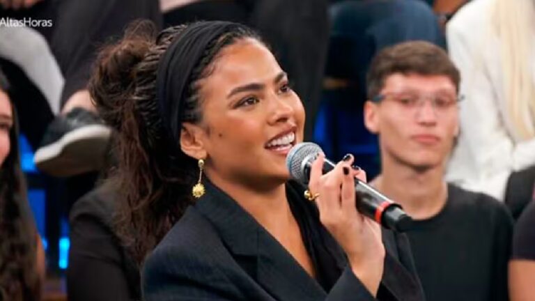 Mulher sorri enquanto segura um microfone e fala em estúdio de televisão com plateia ao fundo