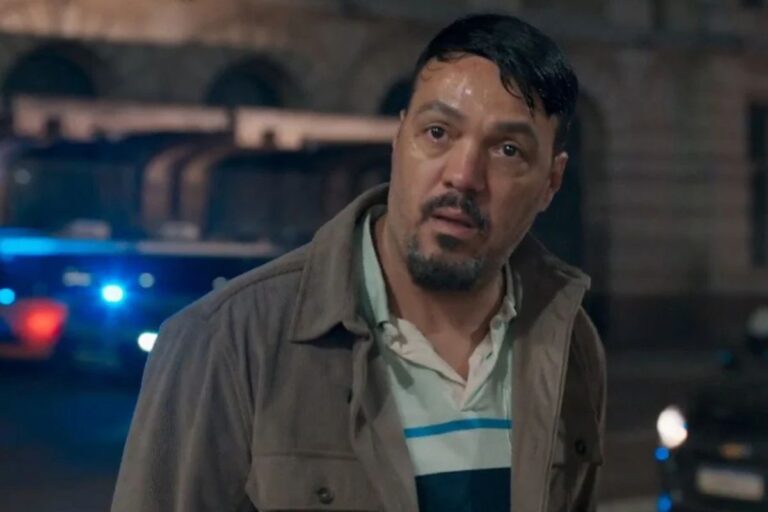 Homem com expressão abatida e rosto suado usa jaqueta marrom em cena noturna com viatura policial e luzes azuis ao fundo