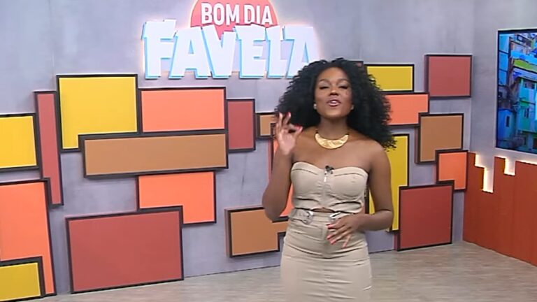 Apresentadora em pé em um estúdio colorido, fazendo um gesto com a mão enquanto sorri; ao fundo, há painéis geométricos em tons quentes e uma placa com o texto “Bom Dia Favela”