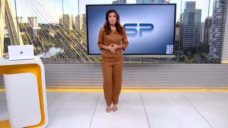 A imagem mostra Sabina Simonato em pé no estúdio do Bom dia SP, telejornal da TV Globo. Ela veste um conjunto marrom e está diante de um telão com o logotipo do programa. Ao fundo, vê-se a paisagem urbana de São Paulo, com destaque para a Ponte Estaiada e prédios modernos, reforçando o cenário característico do noticiário local