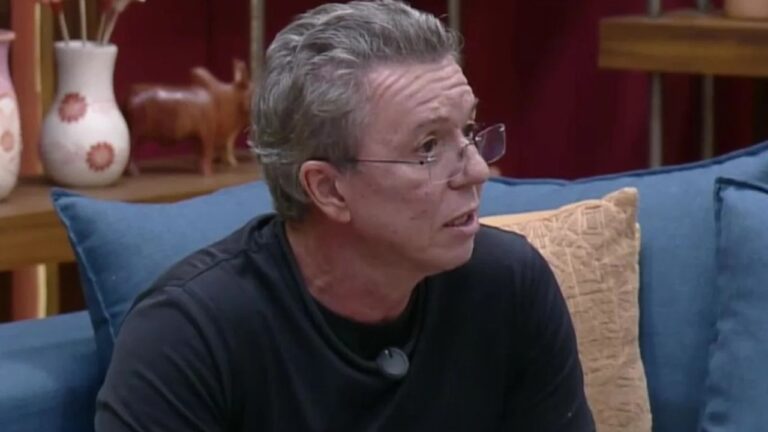 Um homem de meia-idade aparece sentado em um sofá, em ambiente interno que lembra uma sala de estar ou cenário de programa de TV. Ele tem cabelos grisalhos curtos, usa óculos e uma camiseta escura com um microfone preso ao peito. Está de perfil, olhando para a direita, com expressão atenta, como se estivesse ouvindo ou respondendo alguém fora do enquadramento. Ao fundo, há almofadas, uma estante com objetos decorativos e iluminação aconchegante em tons quentes.