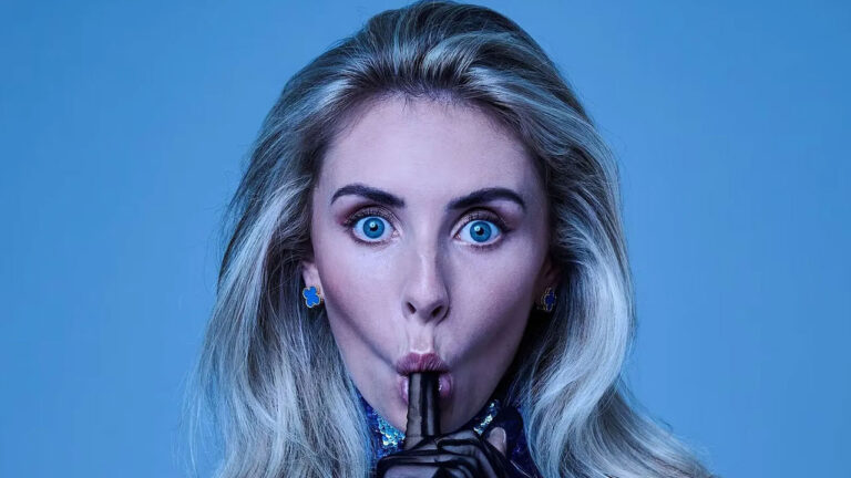 Bonnie Blue encara a câmera com olhos arregalados e dedo indicador na boca usando luvas pretas em frente a um fundo azul claro