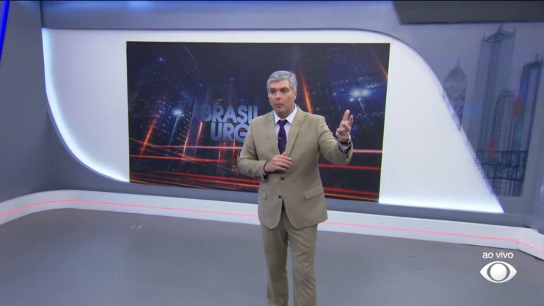 A imagem mostra Joel Datena no estúdio do programa Brasil Urgente, exibido pela Band. Ele usa terno bege e gravata roxa, gesticulando enquanto fala diante de um grande painel com o logotipo do programa e luzes em tons de azul e laranja. No canto inferior direito da tela, aparece o selo ao vivo e o logotipo da emissora