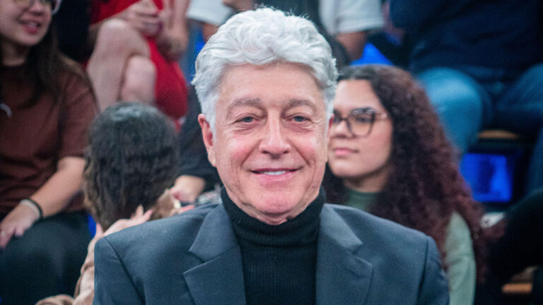 Caco Barcellos sorrindo em primeiro plano com cabelos grisalhos e camisa de gola alta preta, com público ao fundo