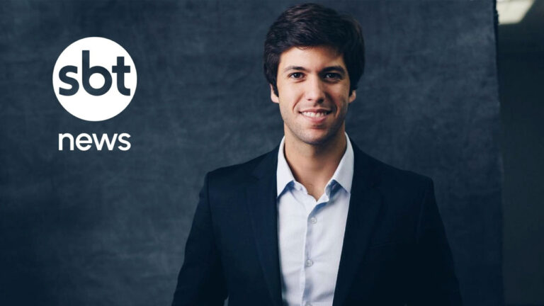 Caio Coppolla sorri usando paletó escuro e camisa clara com o logotipo do SBT News ao fundo