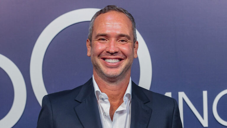Caio Ribeiro sorrindo de terno escuro e camisa branca diante de painel azul com logotipo durante evento da Globo